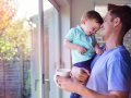 Father holds toddler son while drinking coffee at home, by the window